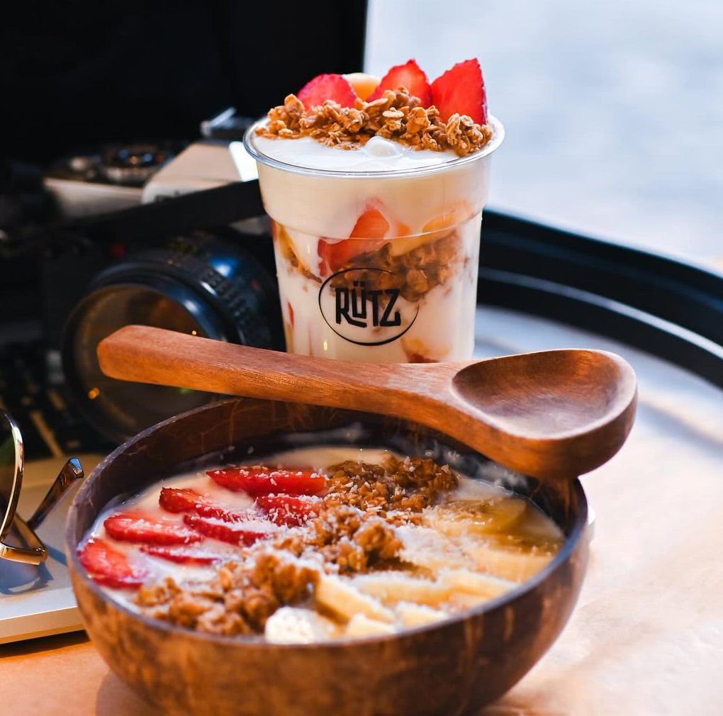 Copo com iogurte, granola e morangos e, à frente, uma tigela de coco com morango, banana, granola e coco ralado, com uma colher de madeira