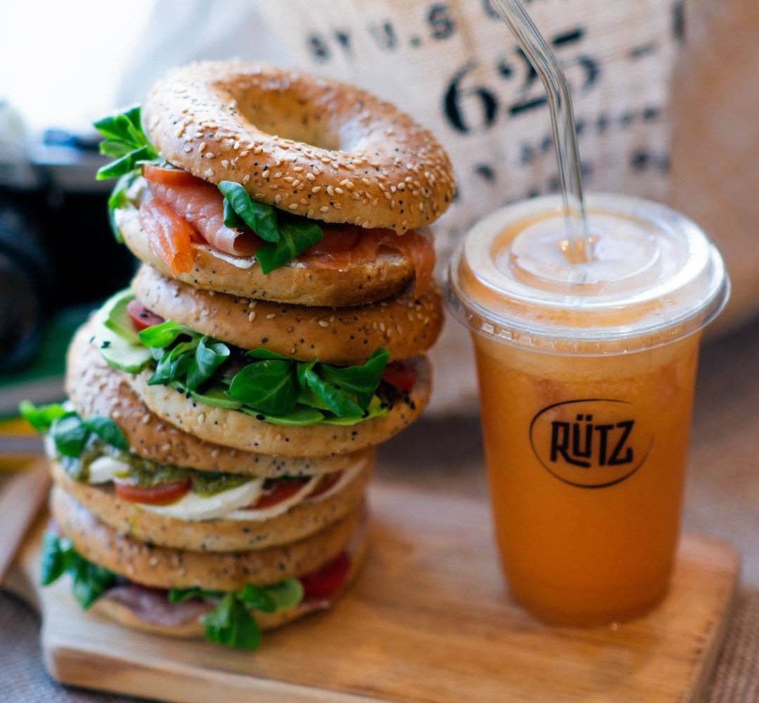 Pilha de bagels com recheios variados, entre os quais salmão e folhas verdes, ao lado de um copo de sumo cor de laranja com palhinha